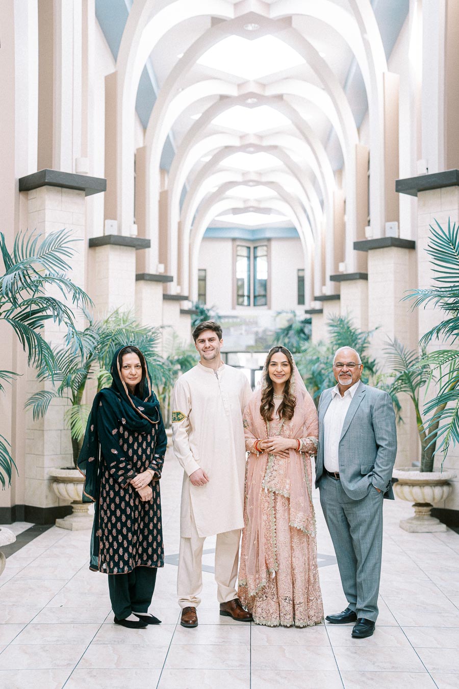 A group of four people wearing traditional and formal attire stands in a bright, elegant hallway with arches and potted plants, smiling at the camera.