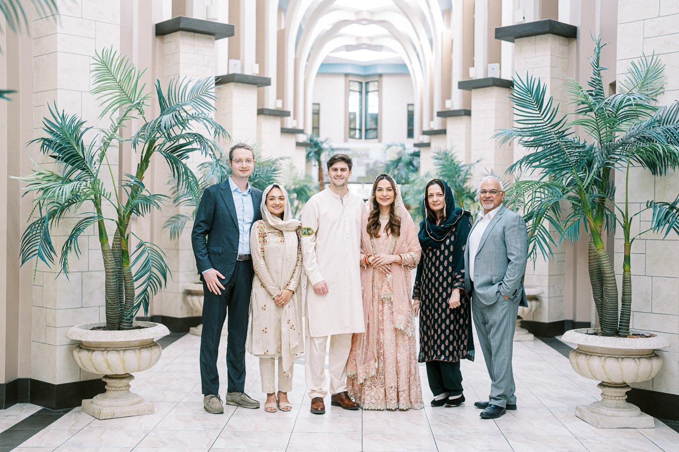 Group of six people in traditional and formal attire standing in a spacious, elegantly designed hallway with potted plants, smiling at the camera.
