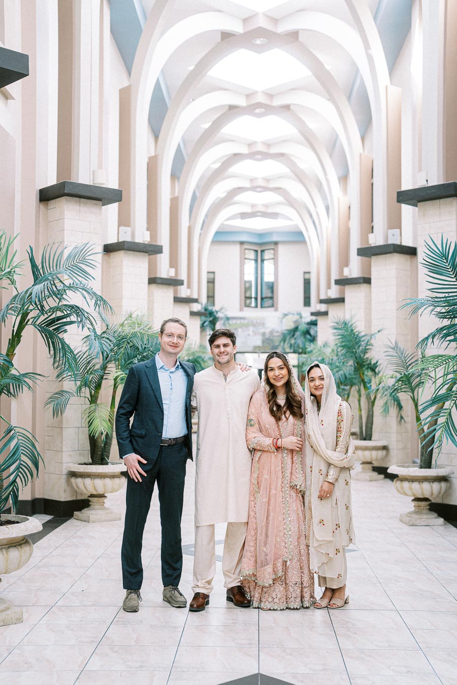 Group of people standing in an elegant, arched hallway adorned with potted plants, wearing a mix of formal and traditional clothing.