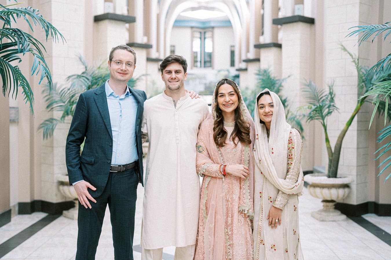A group of four people smiling and posing together indoors, dressed in a mix of traditional and formal attire, surrounded by elegant architecture and plants.