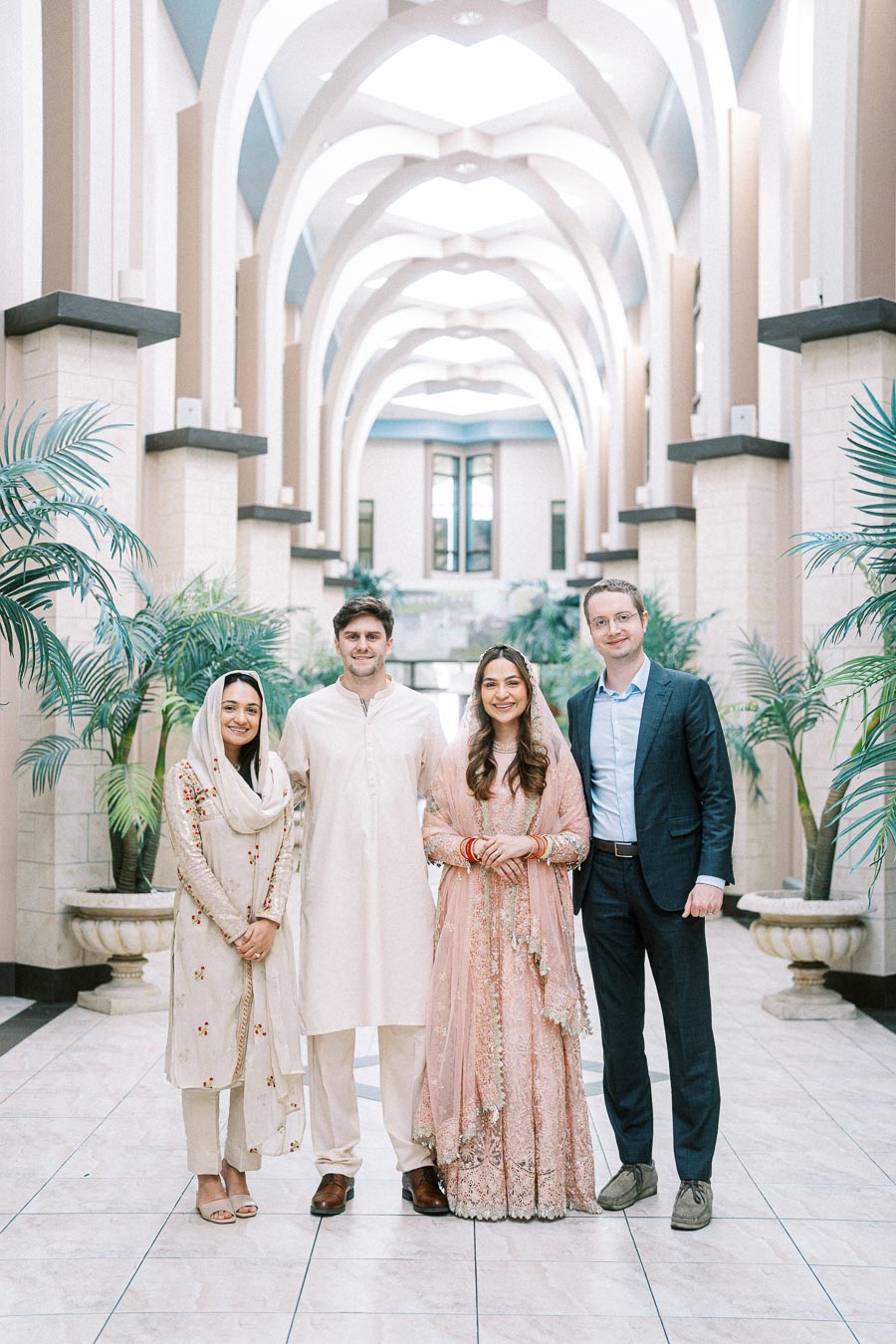 A group of four people posing in a beautifully arched hallway with elegant decor. The women are dressed in traditional Asian attire, featuring intricate embroidery, while the men are wearing formal and traditional outfits. The setting includes tall indoor plants and a bright, spacious atmosphere, suggesting a wedding or cultural event.
