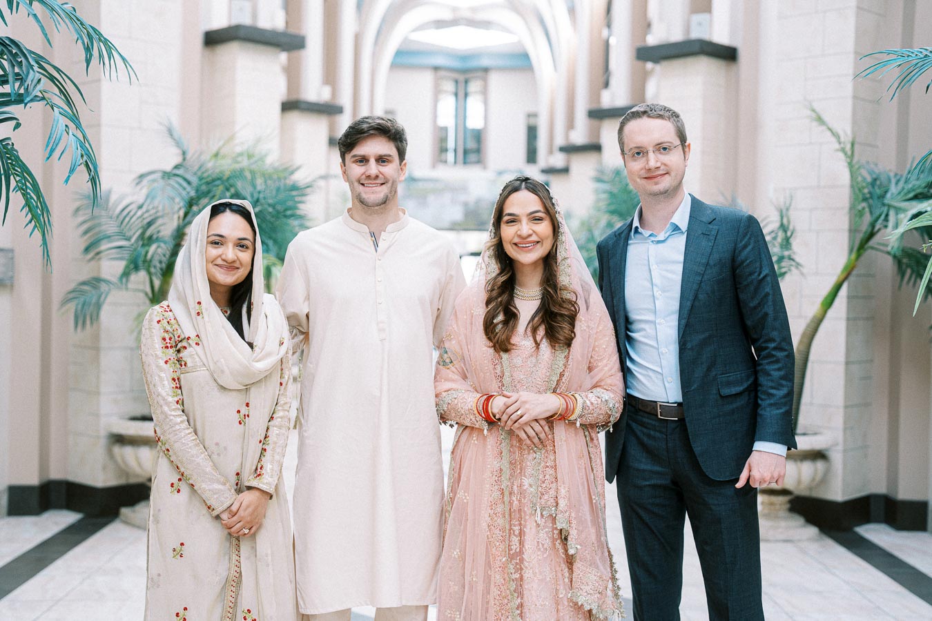 Group of people smiling in an elegant indoor setting with arched architecture and greenery. Two are dressed in traditional attire, and two in modern Western clothing.