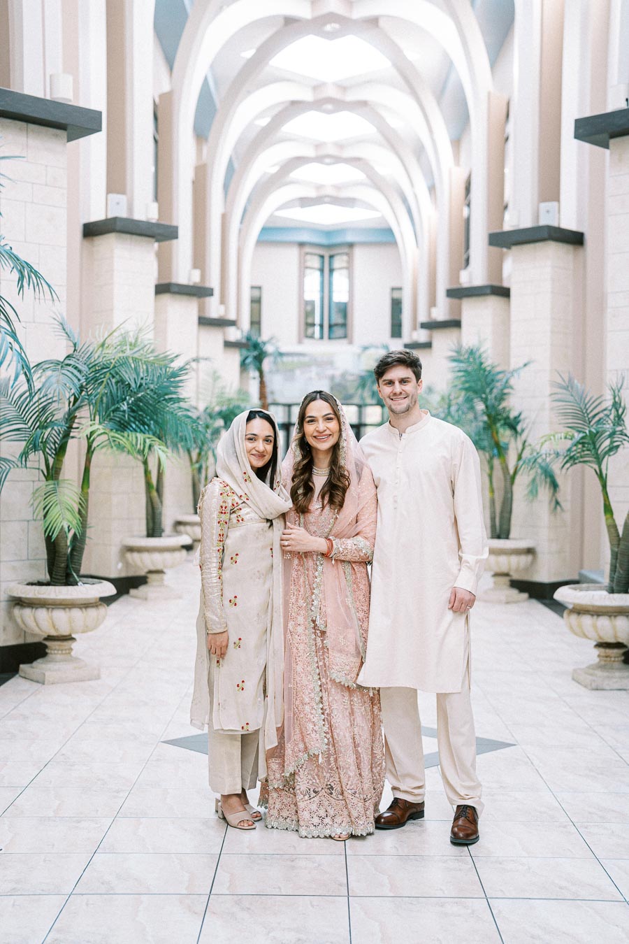 Three people wearing traditional South Asian attire stand smiling in a beautifully decorated interior with tall arches and lush green plants.