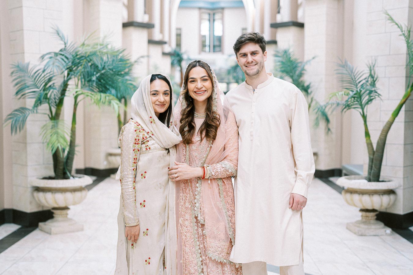 Three people in traditional attire smiling in a beautifully decorated hallway with plants, showcasing cultural elegance and joyful celebration.