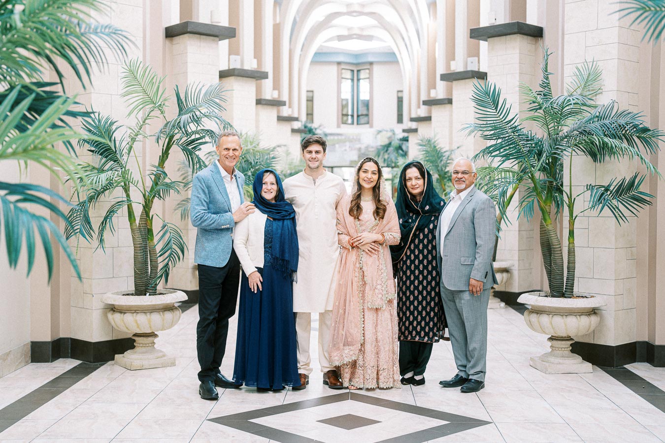 A group of six people dressed in formal attire stands together in a beautifully decorated hallway with potted plants and an arched ceiling. The setting has an elegant and celebratory atmosphere.