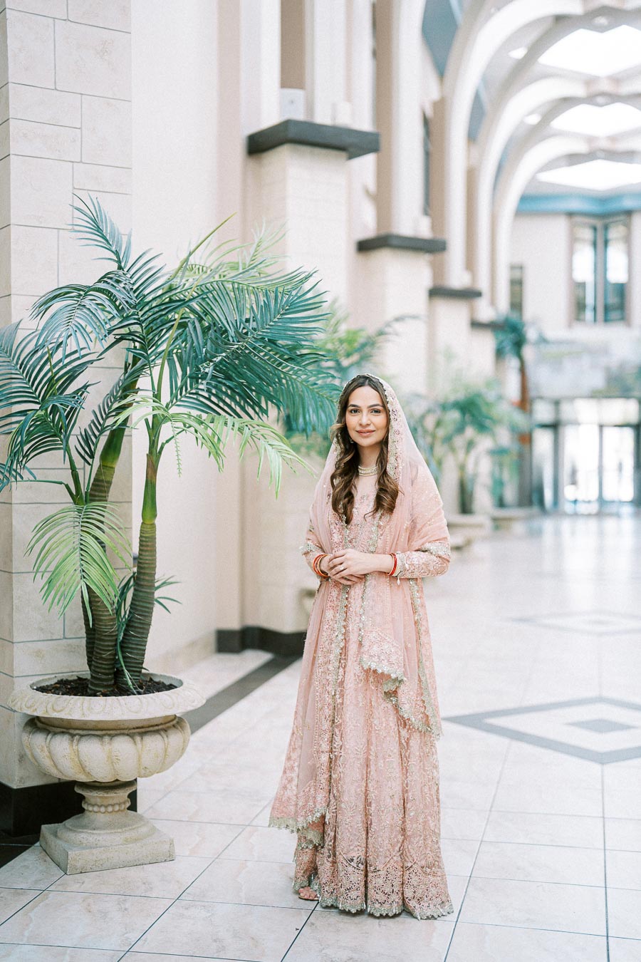 A woman in an elegant pink traditional outfit stands in a spacious, ornate hallway with tall potted plants and arched ceilings, creating a serene and sophisticated atmosphere.