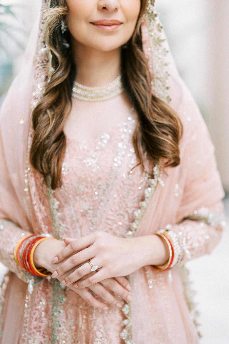 A bride dressed in a pink embellished traditional outfit, with intricate embroidery and beading, paired with bangles and a diamond ring, showcasing elegant bridal fashion.