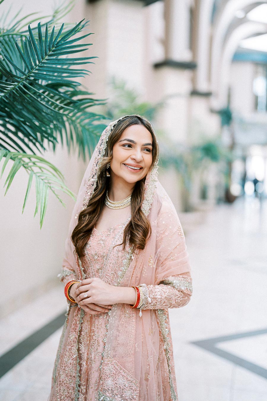 A woman in an elegant pink embroidered traditional dress and dupatta, smiling in a bright, spacious hallway with decorative plants.