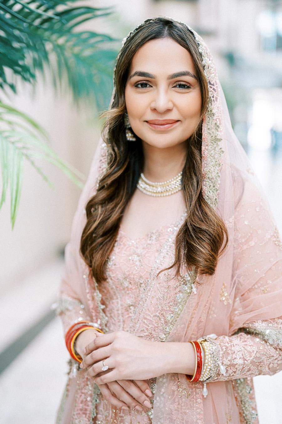 A woman in an elegant pink traditional dress with intricate embroidery, accessorized with red and gold bangles and pearl jewelry, standing in a softly lit setting with green foliage in the background.