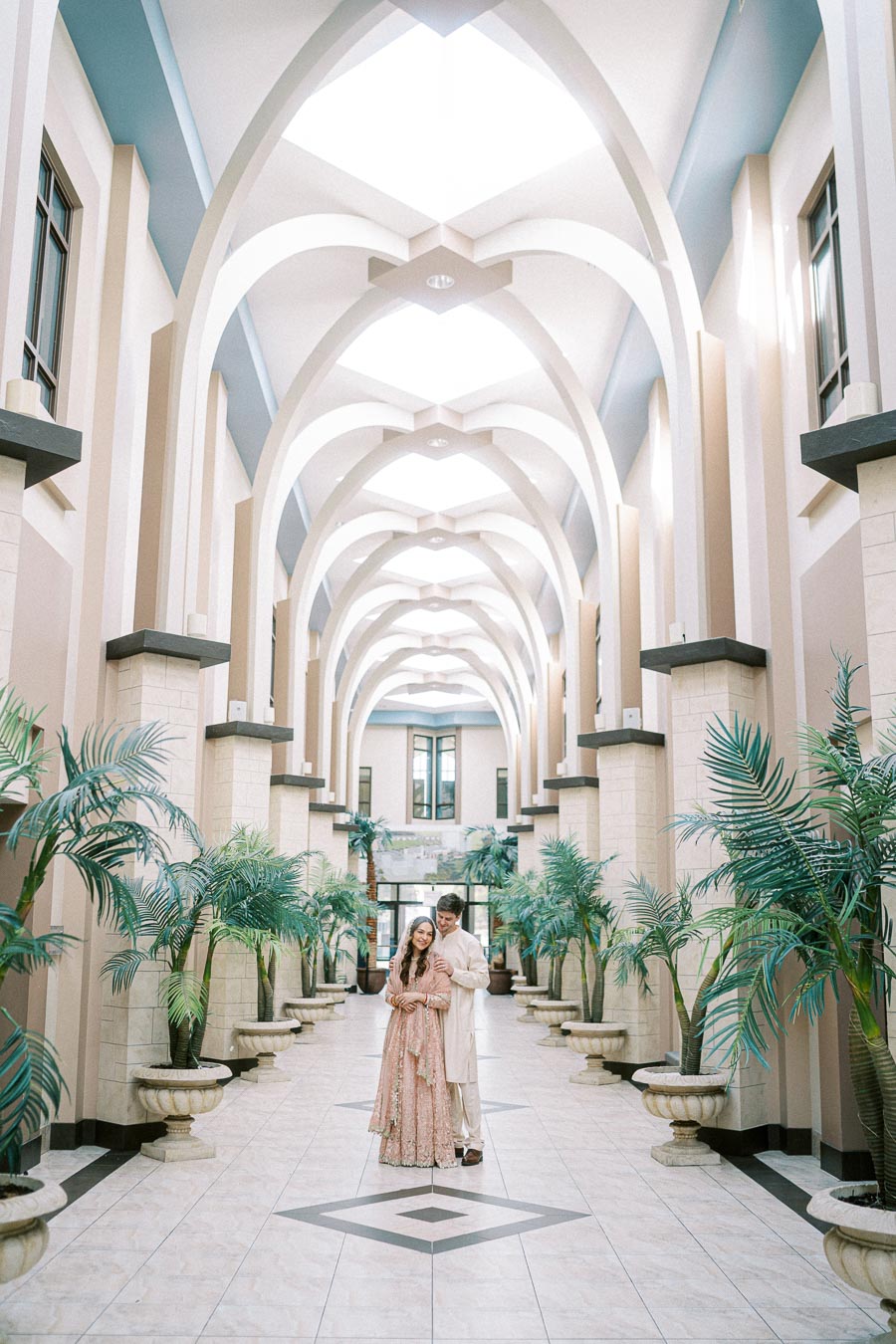 Elegant couple posing in a luxurious hallway with tall arches and decorative plants, showcasing traditional attire.
