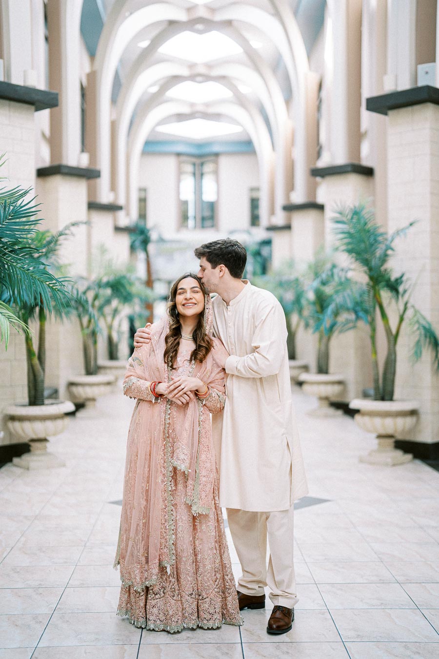 Couple in traditional attire smiling in an elegant hallway with arched ceilings and palm plants, embracing romantic South Asian cultural elements.