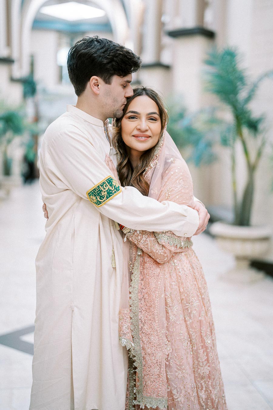 A couple embraces in a beautifully decorated indoor setting, with the woman wearing an embellished pink traditional outfit and the man in a light-colored outfit, capturing a joyful and intimate moment.