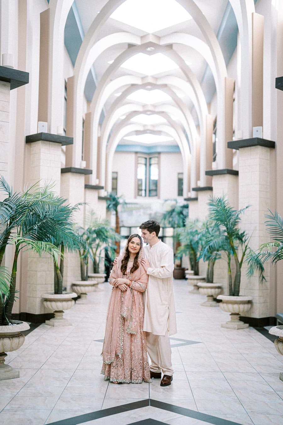 A couple in elegant traditional attire standing in a bright, arched hallway with decorative palm plants and ornate architecture.