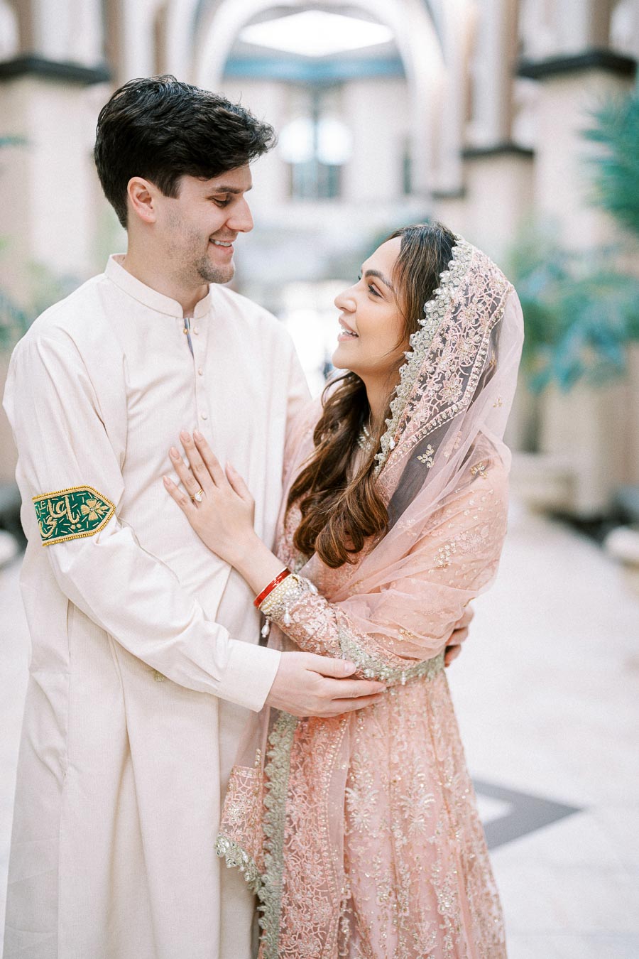 A happy couple in traditional South Asian wedding attire, embracing and smiling at each other in an elegant indoor setting, showcasing cultural richness and love.
