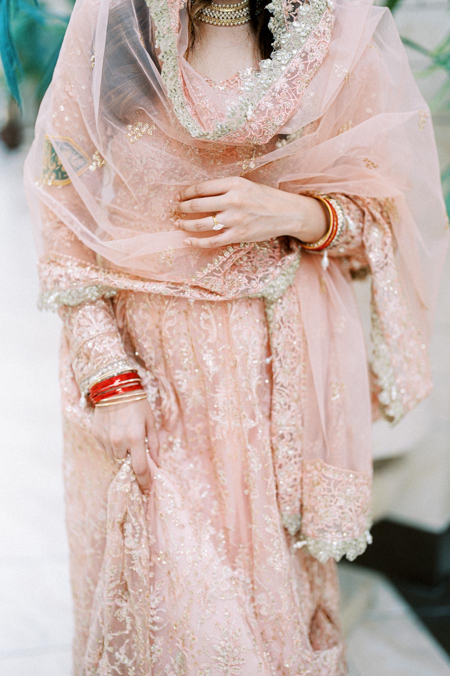 A woman in an intricately designed pink embroidered traditional outfit, adorned with lace and beadwork, holding part of her dress. She wears red bangles and a sparkling ring on her finger, adding elegance and cultural significance to the attire.