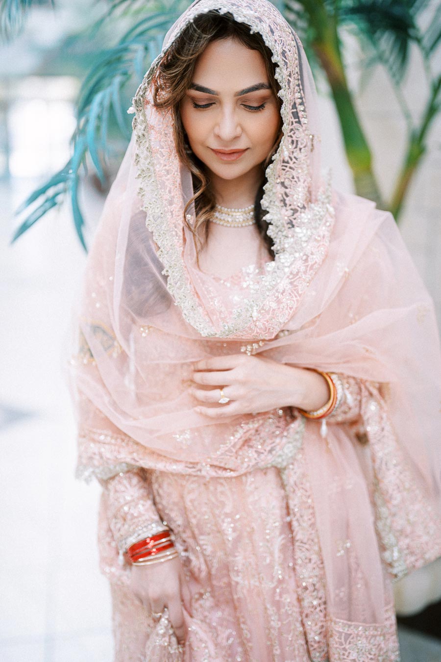 Elegant woman in a pink bridal outfit adorned with intricate embroidery and delicate sequins, wearing traditional red and gold bangles, in a serene setting with greenery in the background.