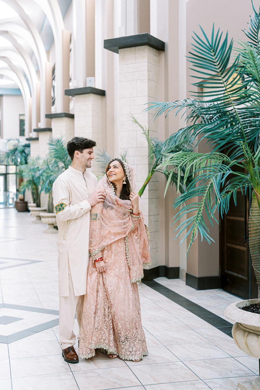 Happy couple dressed in elegant traditional attire, standing in a beautifully decorated hallway with ornate columns and lush green plants.