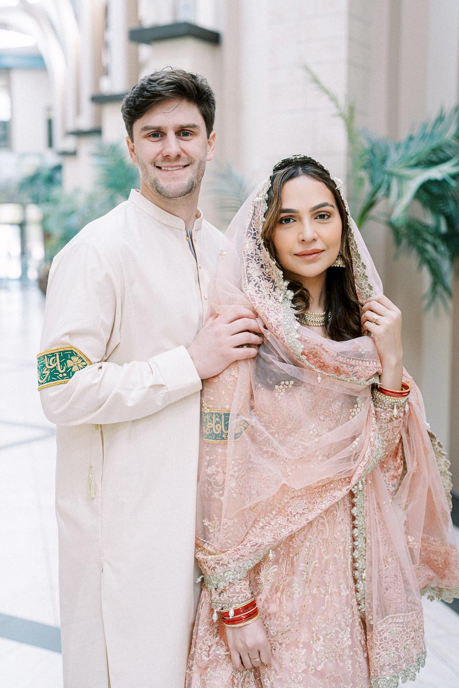 Man and woman in traditional South Asian wedding attire, smiling and posing indoors with elegant architectural details in the background.