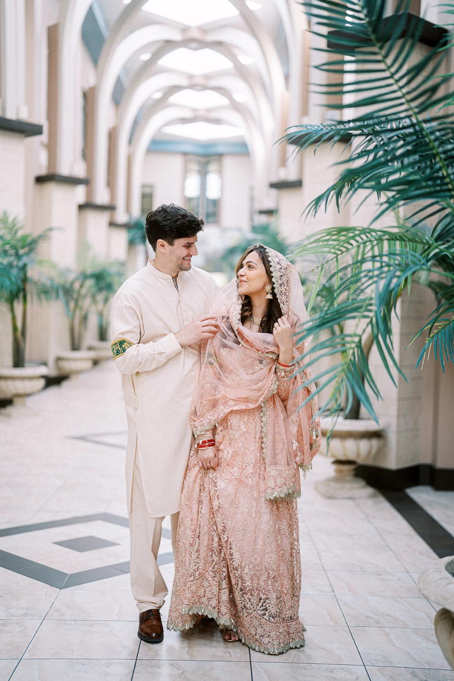 A couple in traditional attire looking at each other, standing in an elegantly designed hallway with arched ceilings and lush green plants.