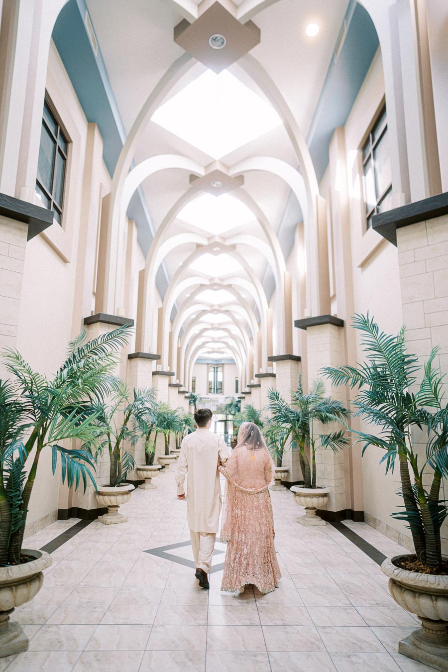 A couple in traditional attire walking hand in hand down an elegant, arched hallway with potted plants and natural light shining through the skylights.