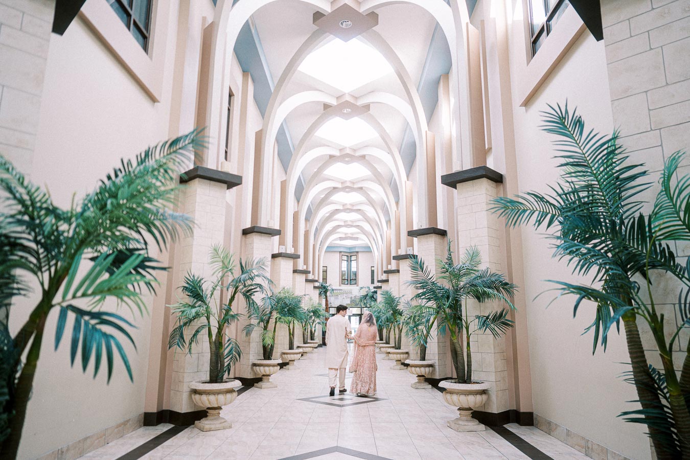 A couple walks through an elegant, arched hallway adorned with lush potted plants, creating a serene atmosphere in a beautifully designed indoor space.
