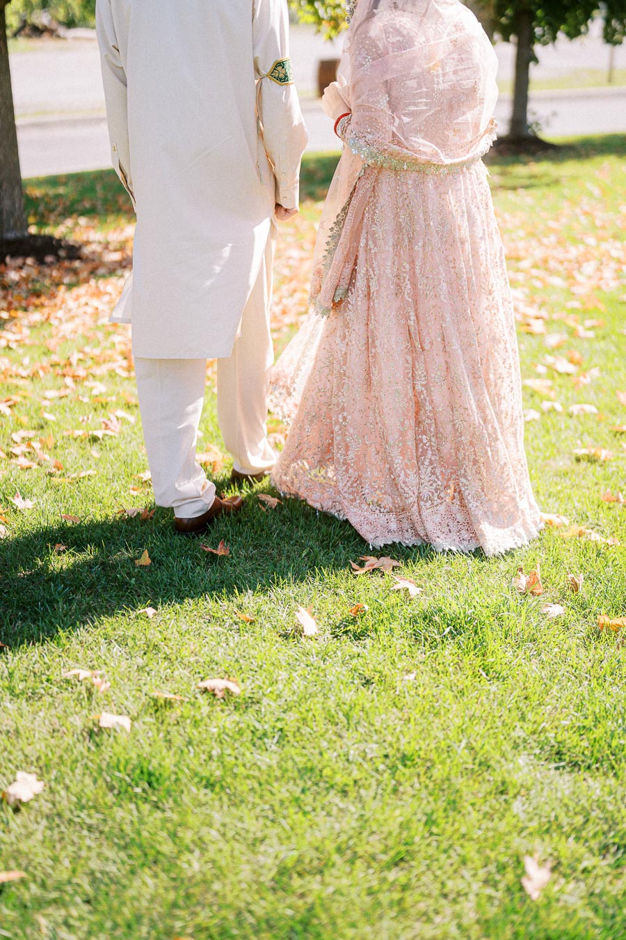 A couple in traditional South Asian attire walk hand in hand on a grassy field, surrounded by scattered autumn leaves, back view.