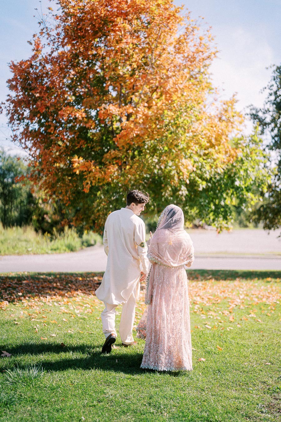 A couple dressed in traditional attire walks hand in hand under a vibrant autumn tree, capturing a serene outdoor moment.