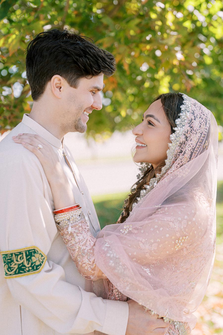 A couple in traditional attire smiling at each other outdoors under a tree, with the woman wearing a pink embroidered shawl.