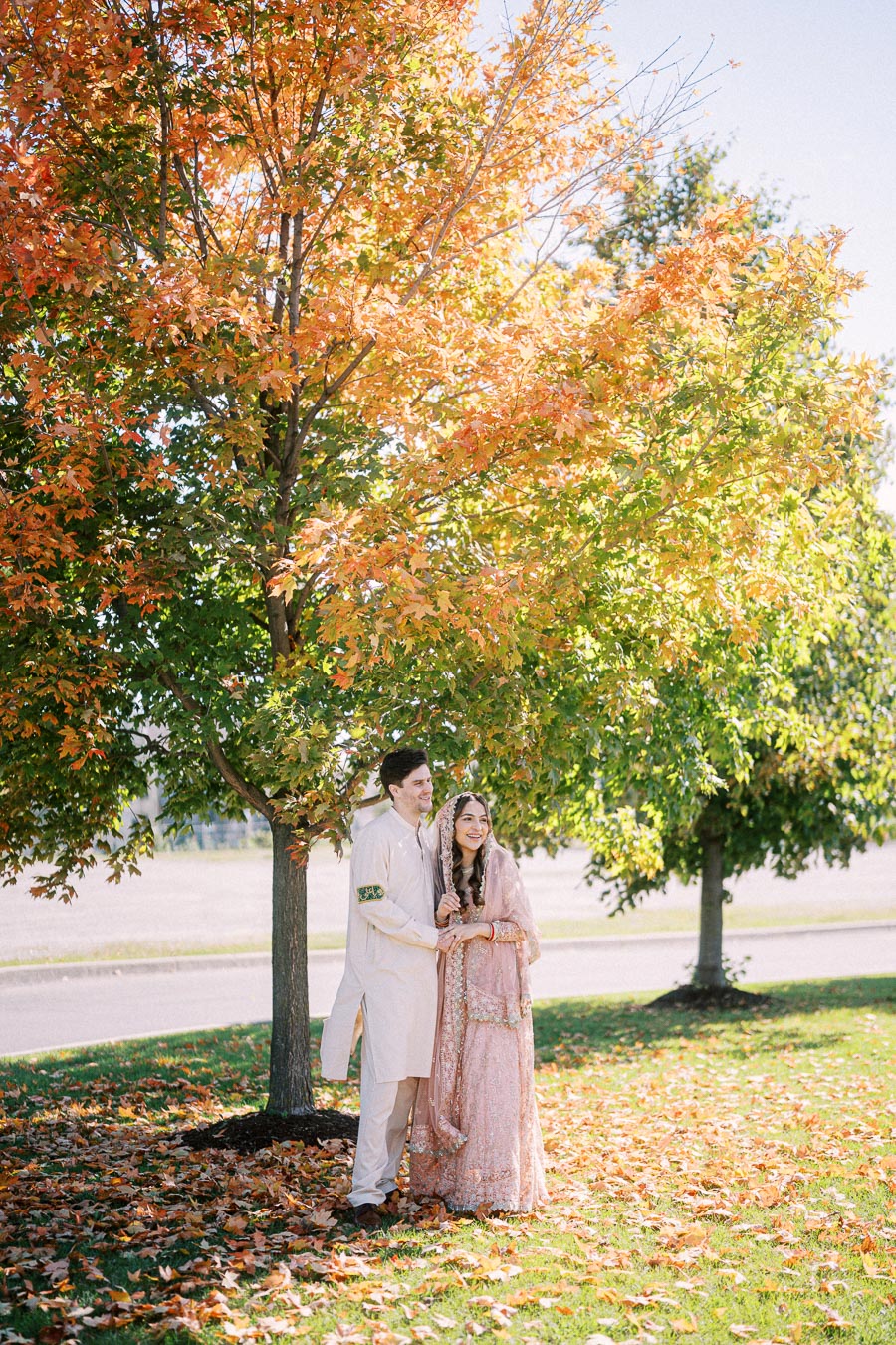 A couple in traditional attire stands under a vibrant autumn tree with colorful leaves, enjoying a sunny day outdoors.