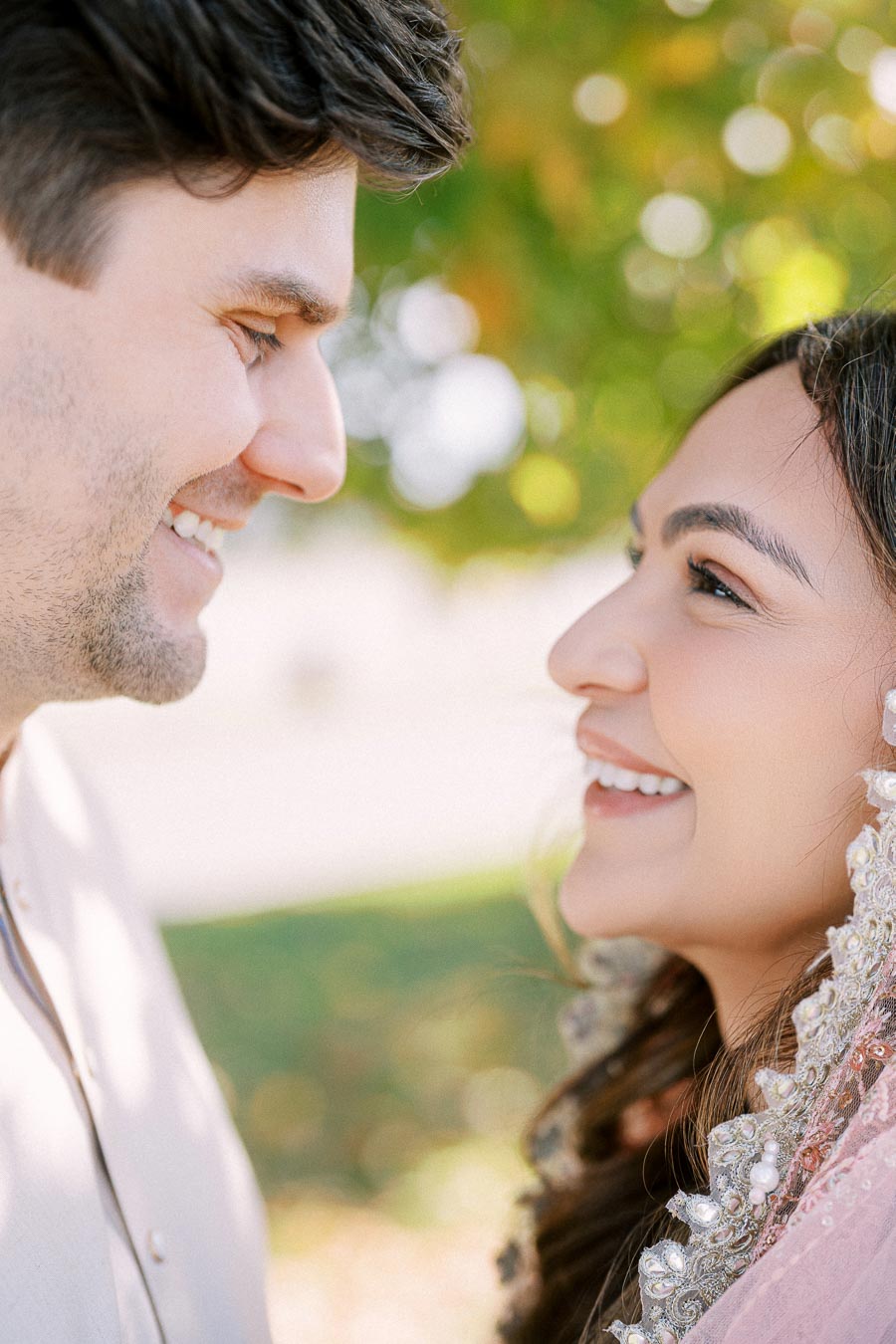 A couple smiling at each other surrounded by a blurred, sunny garden background, highlighting their happiness and close connection.