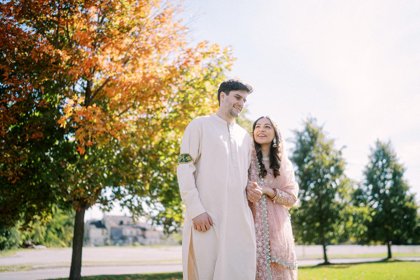 Couple in traditional attire walking outdoors during autumn with vibrant fall foliage in the background, showcasing cultural wedding celebration.