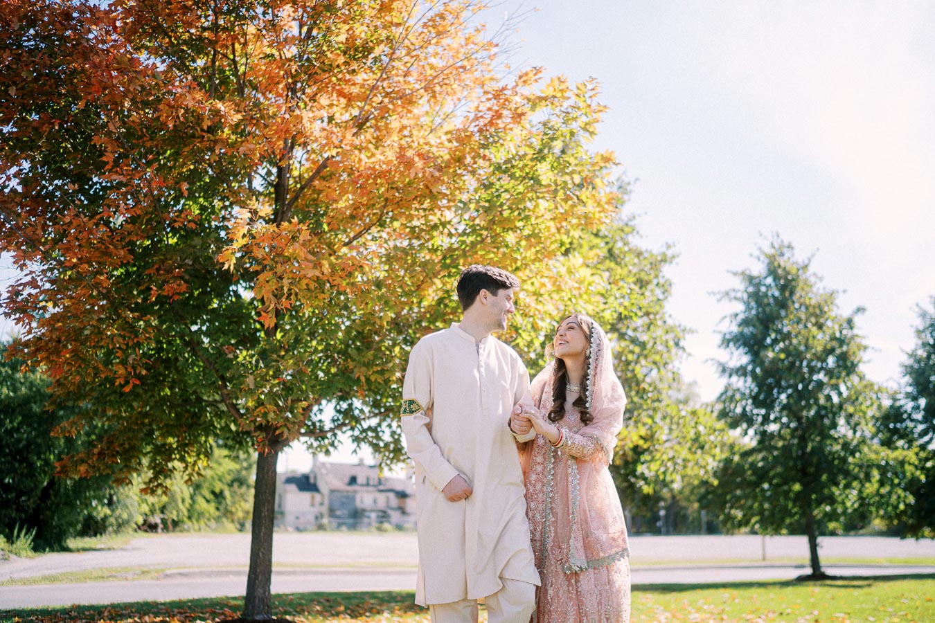 A couple dressed in traditional South Asian attire walking under a tree with autumn leaves in a park, smiling at each other on a sunny day.