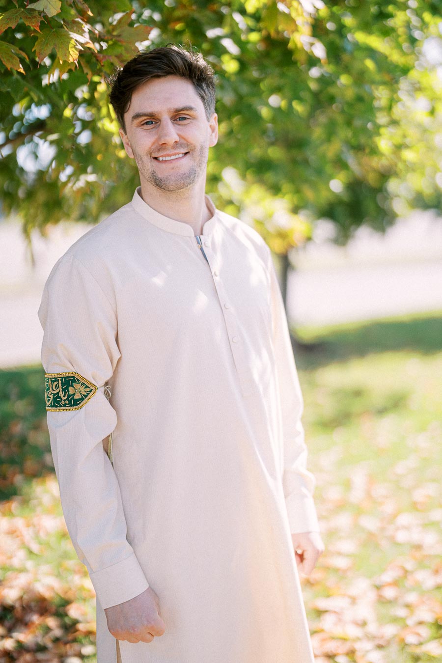 A smiling person in a light-colored traditional outfit stands outdoors under a tree, with sunlight filtering through the leaves.