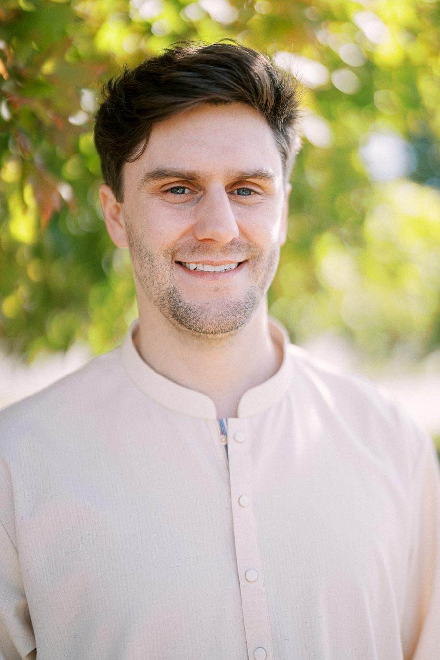 A man wearing a light-colored shirt smiles outdoors with a leafy background.