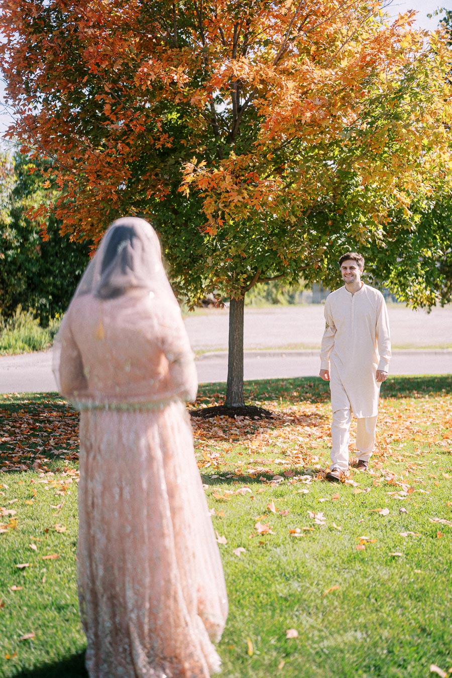 A man in traditional attire walks towards a woman in a bridal gown, standing on a grassy field with autumn leaves and a vibrant tree in the background.