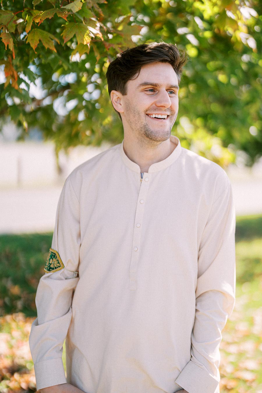 Smiling man wearing a light beige shirt stands outdoors under a green tree with sunlight filtering through the leaves.
