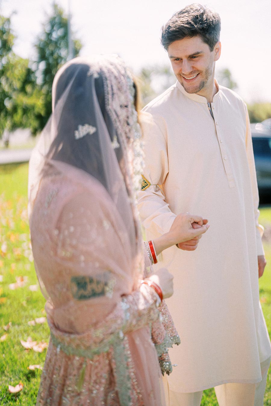 Smiling groom in cream traditional attire lovingly holds hands with bride in embellished pink outfit and veil during outdoor wedding ceremony.