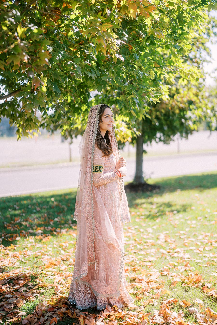 A woman in an elegant, traditional pink dress stands on grass covered with autumn leaves beneath a leafy tree on a sunny day.