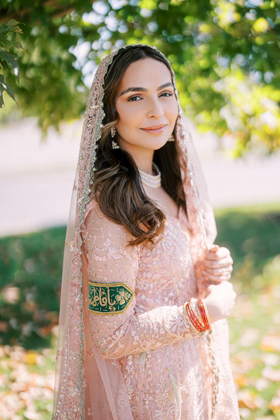 A woman in an elegant pink traditional dress with intricate embroidery, adorned with red bangles and a bejeweled headscarf, standing outdoors with trees and soft sunlight in the background.