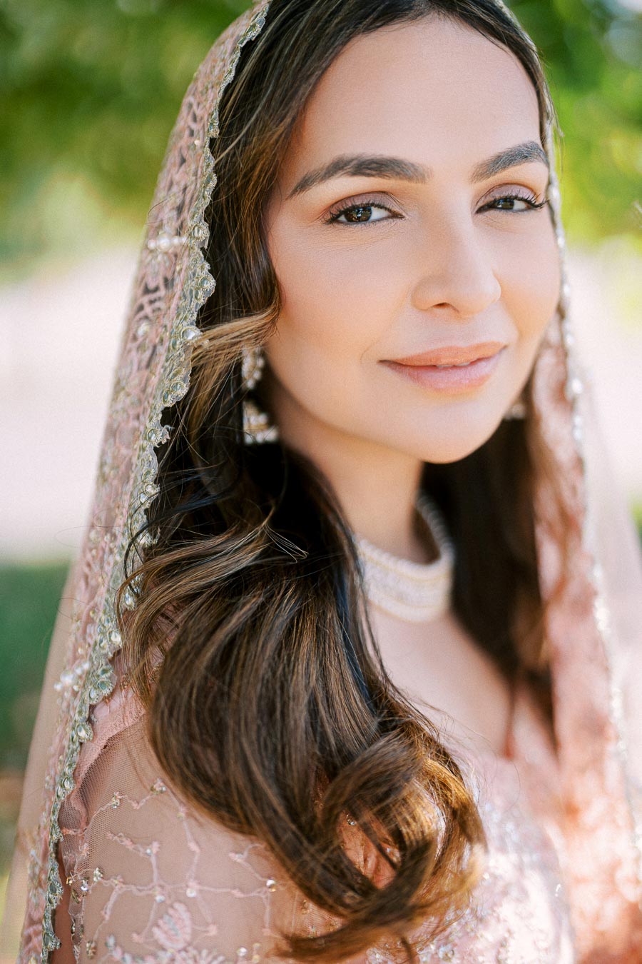 A woman in an elegant pink embroidered traditional outfit, smiling softly, with long wavy hair and a sheer, beaded headscarf, set against a blurred outdoor background with greenery.