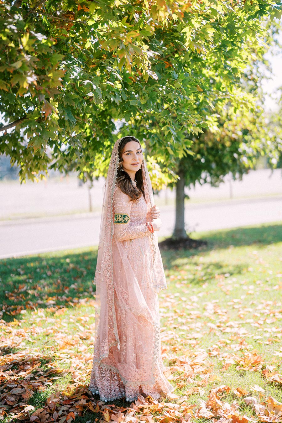 A woman in a delicate pink lace dress stands outdoors under a tree with vibrant green leaves, surrounded by fallen autumn leaves, exuding a serene and elegant vibe.