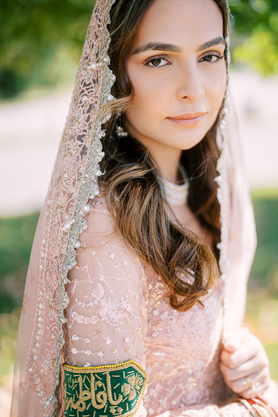 A woman wearing an elegant pink embroidered hijab and dress, standing outdoors with natural light highlighting her soft, wavy hair and intricate lace details.