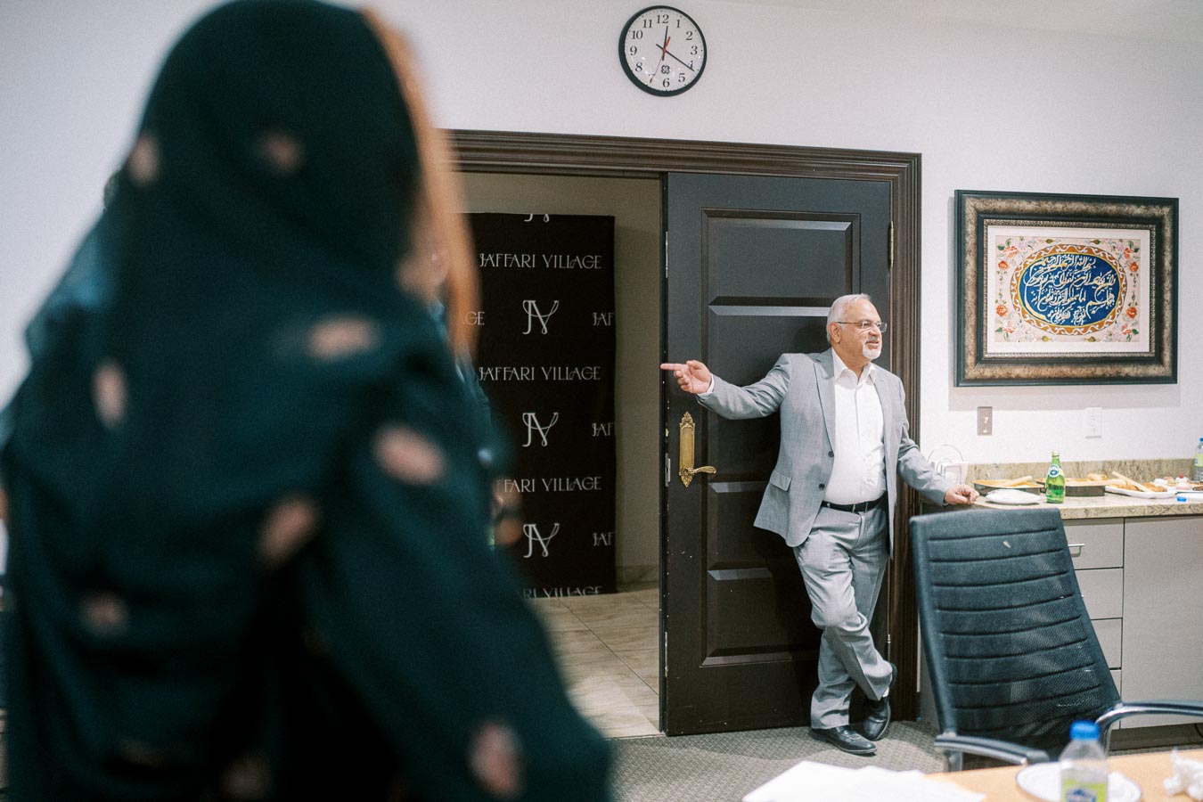 A man in a gray suit stands casually against a door in an office setting, engaging with a woman in a hijab. A wall clock shows the time, and a decorative framed artwork hangs nearby. The room has a conference table with chairs and refreshments.