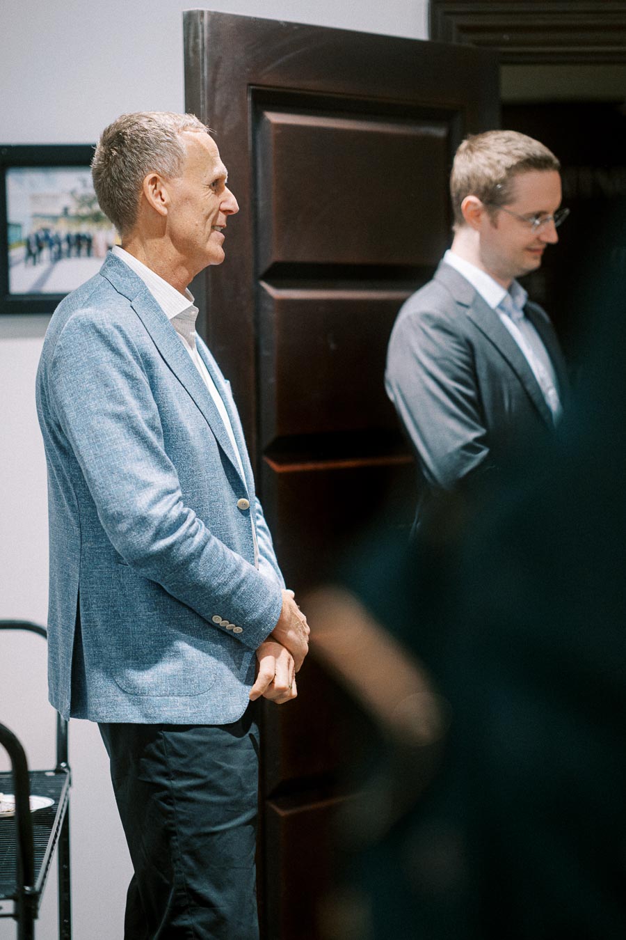 Two men in business attire standing in a room, one wearing a light blue blazer, at a formal event or meeting.