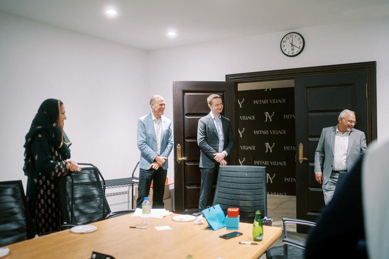 Four people in business attire stand smiling in a conference room, engaged in a meeting. A clock shows the time, and a table with documents and water bottles is in the foreground.