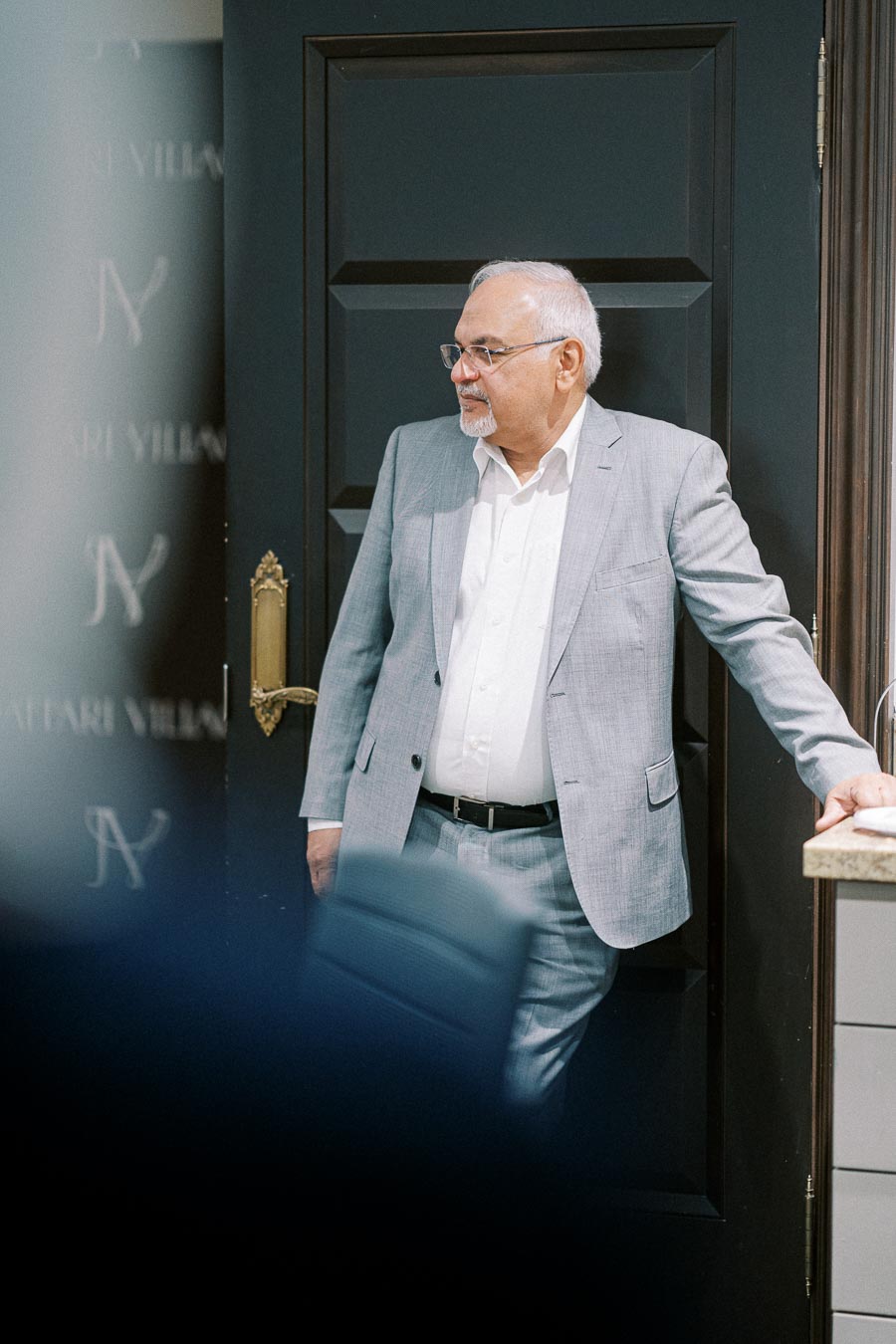 Man in a gray suit leaning against a dark door, looking thoughtful.