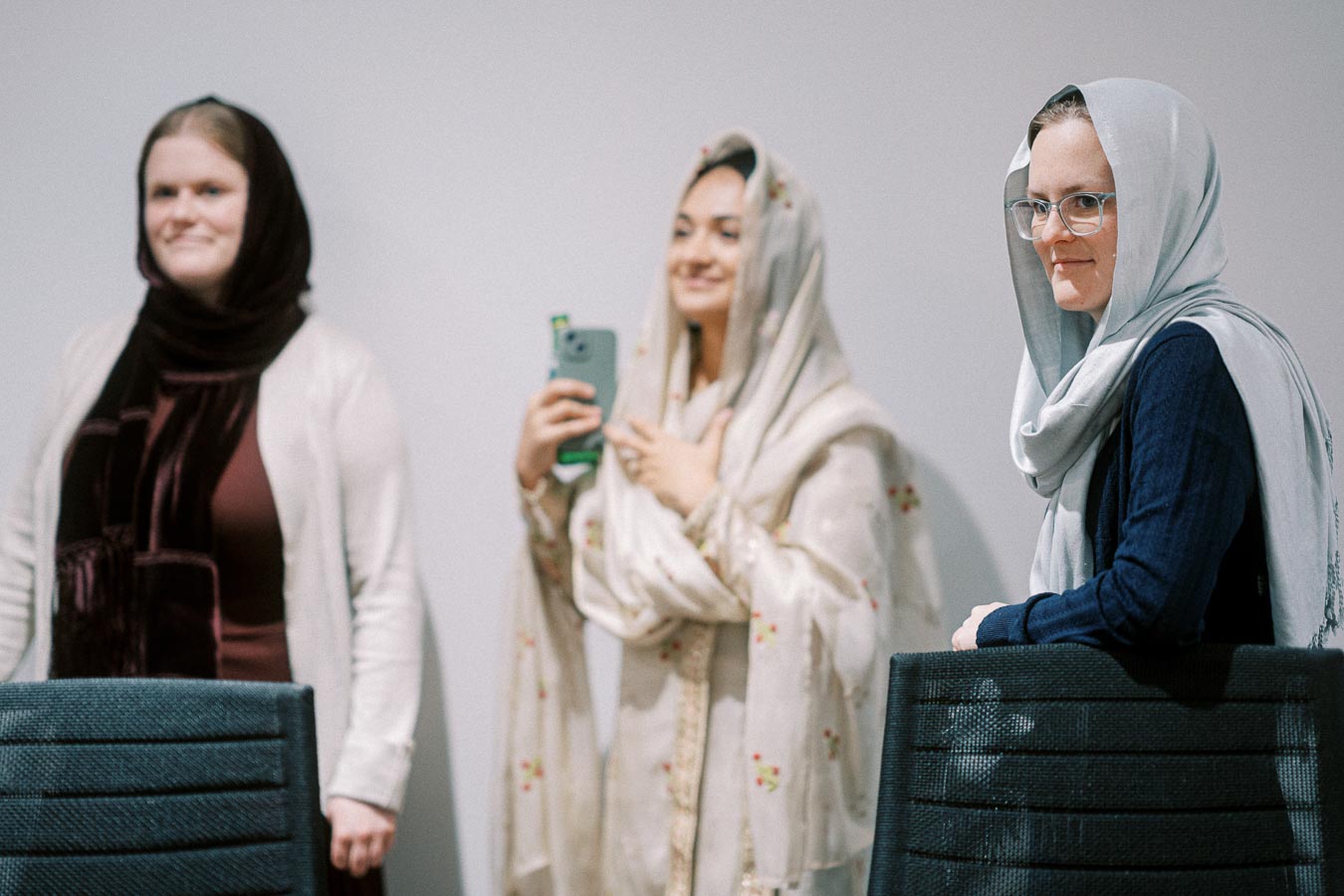 Three women wearing hijabs stand together, facing the camera. One woman in the center is holding a phone and smiling. The setting appears to be indoors, with chairs in front of them.