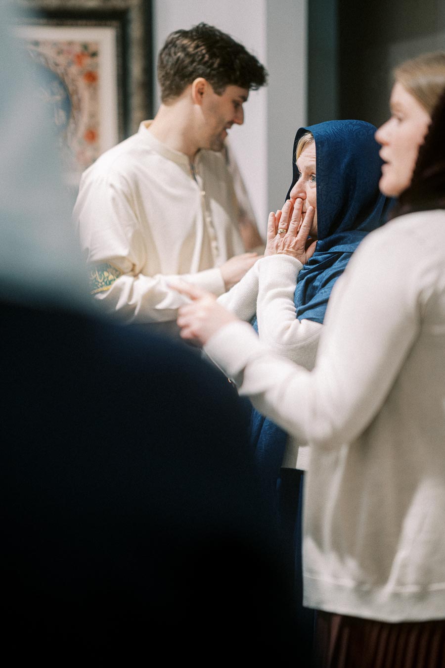 A group of people interacting at an indoor event, with a woman in a blue headscarf looking attentively, suggesting a cultural or social gathering.