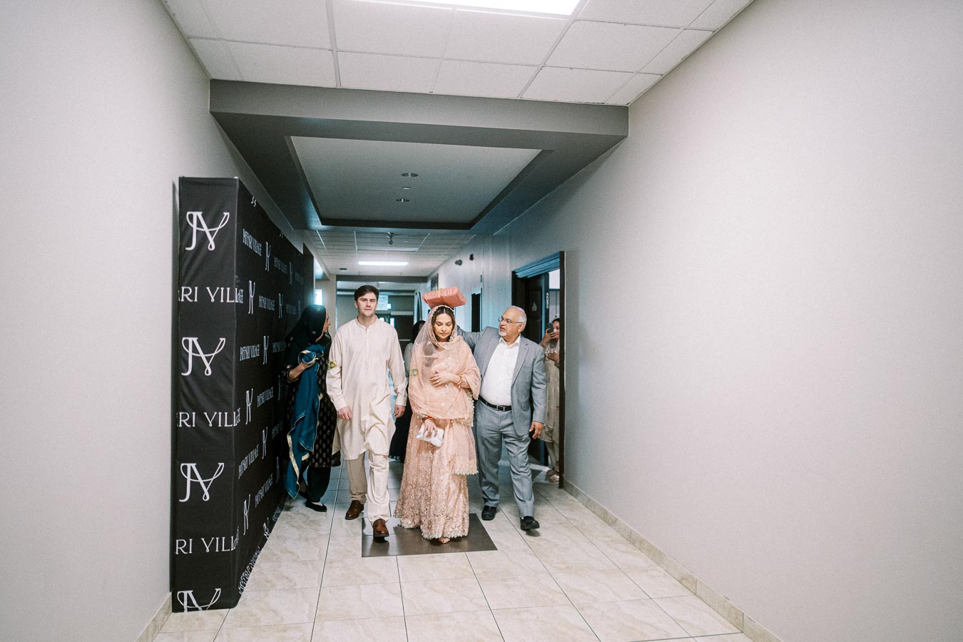 A bride in a peach traditional outfit walks with two men in a hallway decorated with branding banners, preparing for a wedding ceremony.