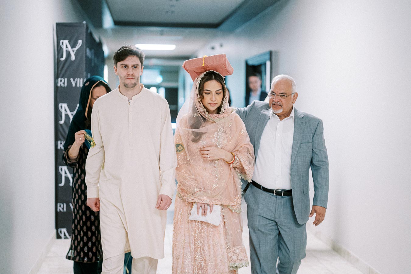 A bride in a traditional pink outfit is walking down a hallway with two men and a woman. The setting appears formal, with one man in traditional attire and the other in a suit, suggesting a wedding or cultural ceremony.